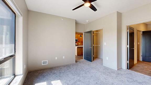 Bright apartment bedroom with soft carpeting, a large window, ceiling fan, neutral walls, and an open doorway leading toward the kitchen and hallway.