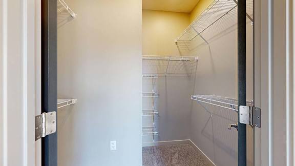 Walk-in apartment closet with carpeted flooring, multiple wire shelving units, and ample storage space for clothing and household items.