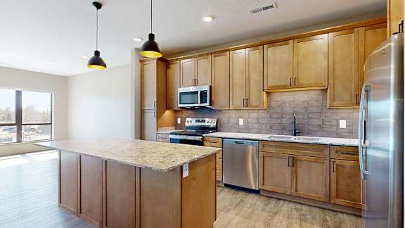 Spacious apartment kitchen with wood cabinetry, a large island, tile backsplash, stainless steel appliances, pendant lighting, and an open layout with adjacent living space and large windows.