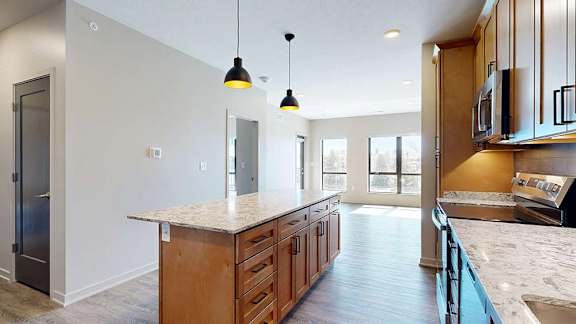 Spacious apartment kitchen with a large island, wood cabinetry, stainless steel appliances, pendant lighting, stone-look countertops, and an open layout flowing into the bright living area.