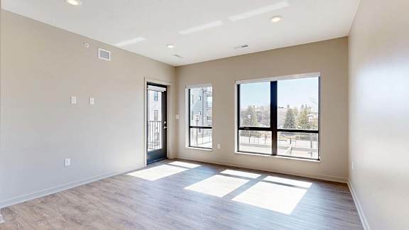 Bright open-concept apartment living area with wood-style flooring, large windows, abundant natural light, and a glass door leading to a private balcony.