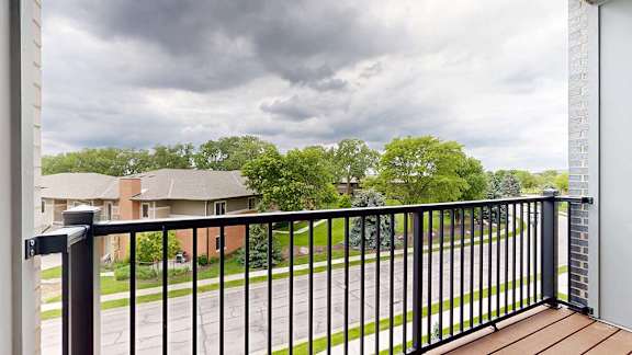 Private apartment balcony with black metal railing and views of trees and neighboring homes under a cloudy sky.