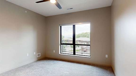 Bright apartment bedroom with carpeted flooring, a large window, and a ceiling fan.