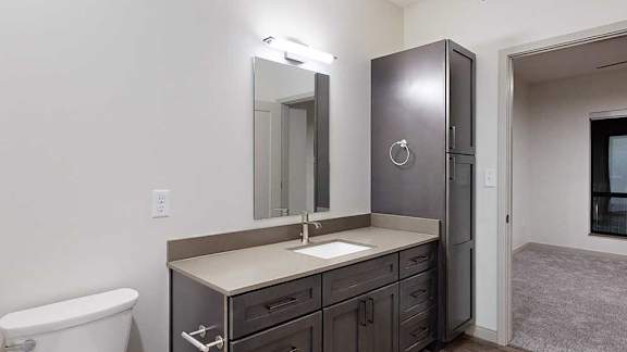Apartment bathroom vanity with dark cabinetry, a light countertop, large mirror, bright lighting, and adjacent bedroom access.