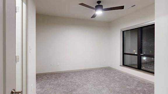 Bright apartment bedroom with neutral carpeting, a ceiling fan, and a large window overlooking nighttime views.