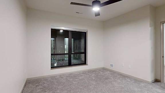 Carpeted apartment bedroom with neutral walls, a ceiling fan, and a large window providing natural light.