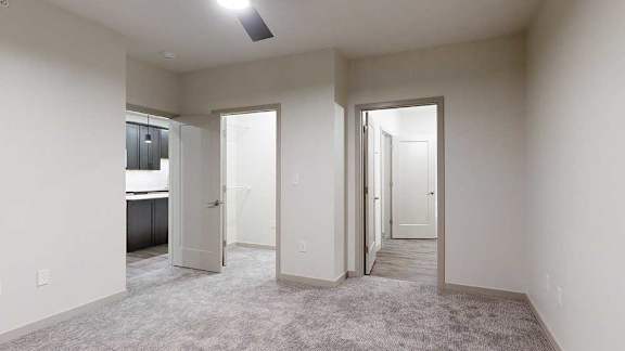 Carpeted apartment bedroom with neutral walls, a ceiling fan, and doorways leading to a bathroom and hallway.
