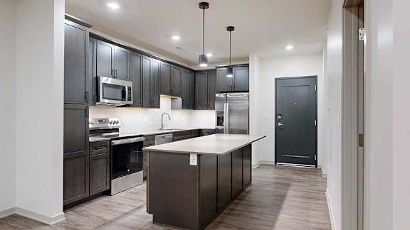 Modern apartment kitchen with dark cabinetry, stainless steel appliances, a center island, pendant lights, and wood-look flooring.
