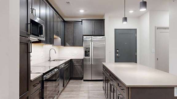 Modern apartment kitchen with dark cabinetry, stainless steel appliances, light countertops, and a large island with pendant lighting.