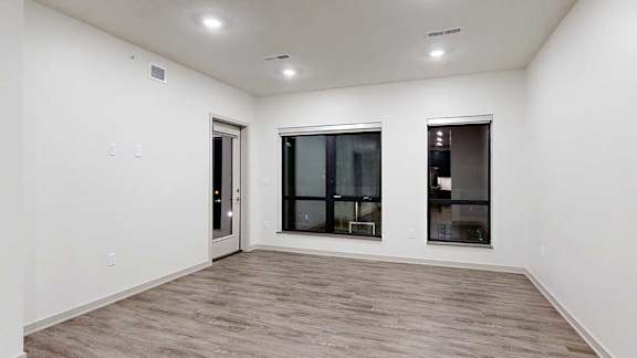 Open apartment living area with wood-look flooring, recessed lighting, large windows, and a glass patio door.