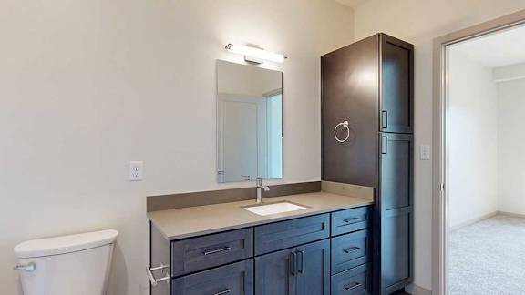 A bathroom with a quartz vanity, dark wooden cabinets, a tall storage cabinet, and a door open to a bedroom.