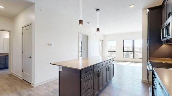 A kitchen with dark wooden cabinets and a large island, in front of a spacious living room with floor-to-ceiling windows.