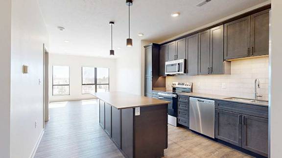A kitchen with dark wooden cabinets and a large island, in front of a spacious living room with floor-to-ceiling windows.