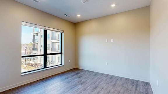Bright apartment room with a large window, wood-style flooring, and natural light.