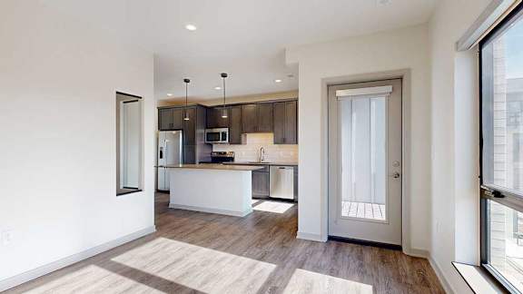 A living room with floor-to-ceiling windows open to a kitchen with a large island with dark cabinets.
