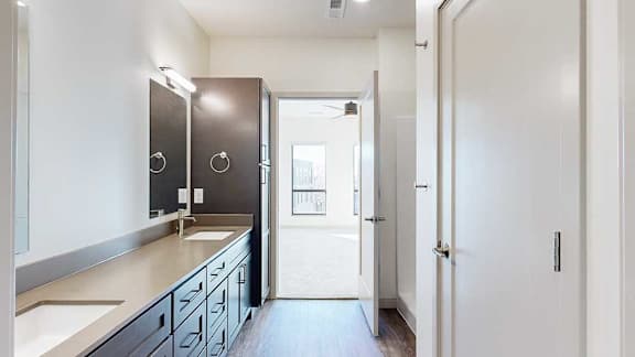 a bathroom with a double quartz vanity sink, two mirrors, and a door to a bedroom