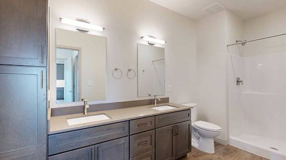 A bathroom with dark wooden cabinets, a quartz double vanity sink, two mirrors with overhead lighting, and tall storage cabinet.