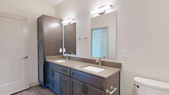 A bathroom with dark wooden cabinets, a quartz double vanity sink, two mirrors with overhead lighting, and tall storage cabinet.