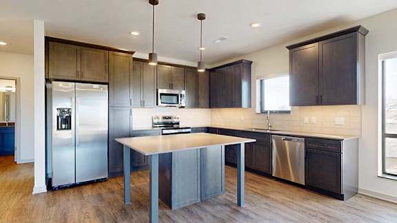 A kitchen with dark wooden cabinets, quartz countertops, a tile backsplash, an island, and a window over the kitchen sink.