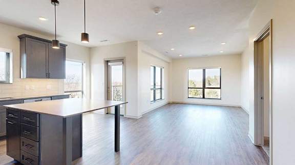 A kitchen with dark wooden cabinets and a large island, in front of a spacious living room with floor-to-ceiling windows.