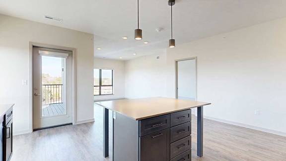 A kitchen with dark wooden cabinets and a large island, in front of a spacious living room with floor-to-ceiling windows.