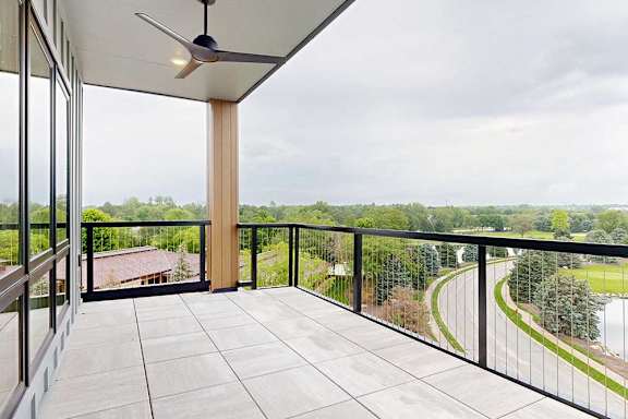 An oversized balcony with stone tile flooring, a metal railing, a ceiling fan, and a stunning view of the neighborhood.