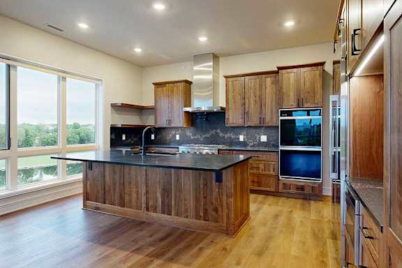 A gourmet kitchen with an oversized island, oak wooden cabinetry, stainless steel commercial appliances, dark quartz countertops, a range hood, and a tile backsplash.