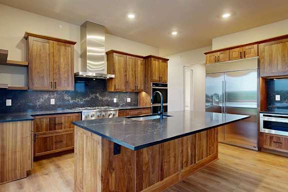 A gourmet kitchen with an oversized island, oak wooden cabinetry, stainless steel commercial appliances, dark quartz countertops, a range hood, and a tile backsplash.