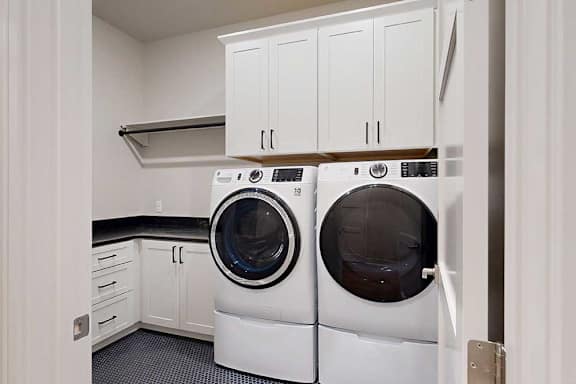 A laundry room with a full-sized front-loading washer and dryer with overhead cabinetry for storage, and a utility sink with quartz counters.