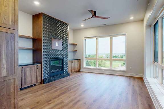 A modern living room with a fire place in a floor-to-ceiling mantel, built-in cabinetry, floor-to-ceiling windows, hardwood-style flooring, and a ceiling fan.