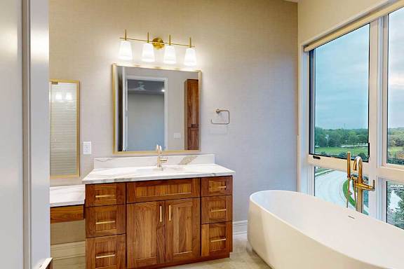 A spacious ensuite bathroom with a double vanity sink, floor-to-ceiling window, soaking tub, and tiled flooring.
