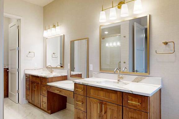 A spacious ensuite bathroom featuring a light quartz double vanity with oak cabinetry, three framed mirrors, overhead lighting, and tiled flooring.
