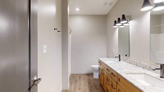 A bathroom with warm wooden cabinets, a quartz double vanity sink, two mirrors with overhead lighting, and tall storage cabinet.