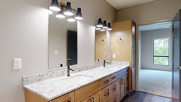 A bathroom with warm wooden cabinets, a quartz double vanity sink, two mirrors with overhead lighting, and tall storage cabinet.