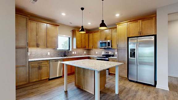 A kitchen with warm wooden cabinets, quartz countertops, an island, and stainless steel appliances.