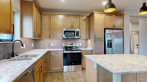 A kitchen with warm wooden cabinets, quartz countertops, an island, and stainless steel appliances.