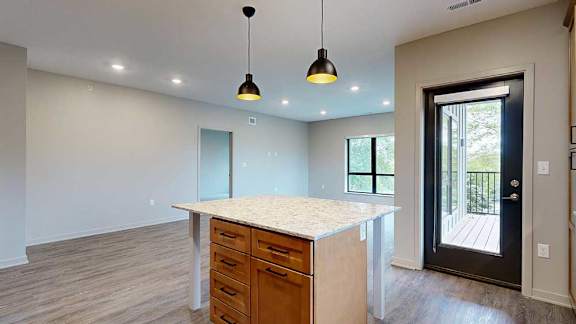 A kitchen with warm wooden cabinets and a large island, in front of a spacious living room with floor-to-ceiling windows.