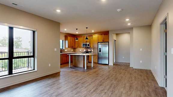 A living room with floor-to-ceiling windows open to a kitchen with a large island with warm wooden cabinets.