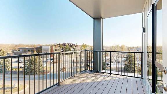A balcony with a black railing and wooden floor overlooks a parking lot and buildings.