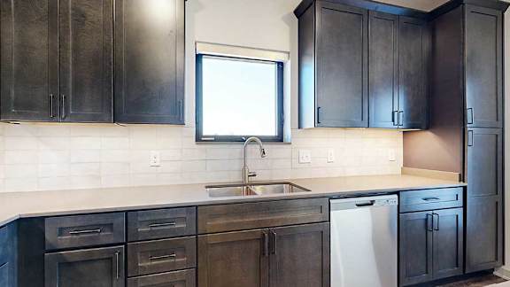 a kitchen with dark wood cabinets, a stainless steel dishwasher and sink, a tile backsplash, and a window above the sink