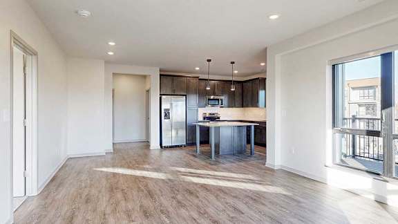 a living room with a large window and hard wood flooring in front of a modern kitchen with dark cabinets