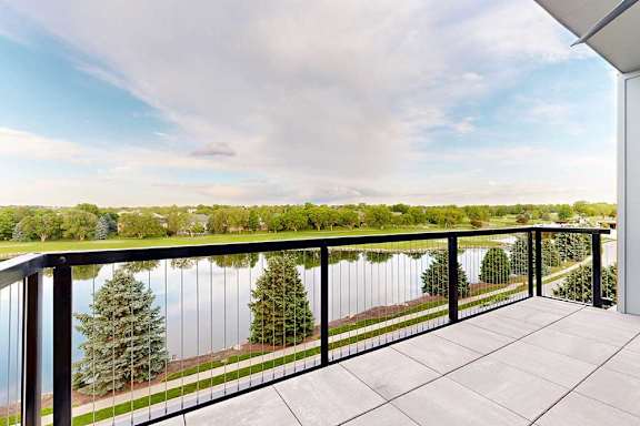 A large balcony in a penthouse apartment overlooks a body of water with trees in the distance.