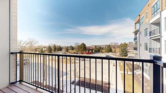 Private apartment balcony with black metal railing overlooking a courtyard, neighboring buildings, trees, and open blue sky.