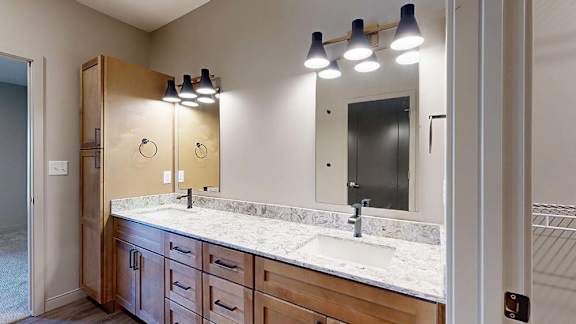 Modern apartment bathroom with a double-sink vanity, wood cabinetry, quartz-style countertop, large mirror, and contemporary wall-mounted lighting.