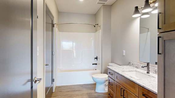 Modern apartment bathroom with a quartz-style vanity, wood cabinetry, a large mirror, and a tub-shower combination with a sliding glass shower door.