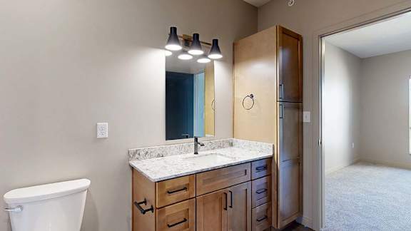 Modern apartment bathroom with a quartz-style vanity, wood cabinetry, contemporary lighting, and a built-in linen cabinet, with the adjacent bedroom visible through the doorway.