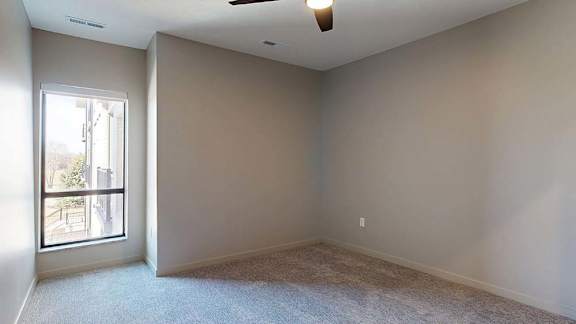 Bright apartment bedroom with neutral carpeting, recessed lighting, and a large window that brings in natural light.