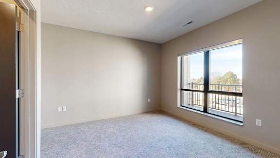 Bright apartment bedroom with neutral carpeting, a large window overlooking the balcony, and soft natural light creating an open, airy feel.