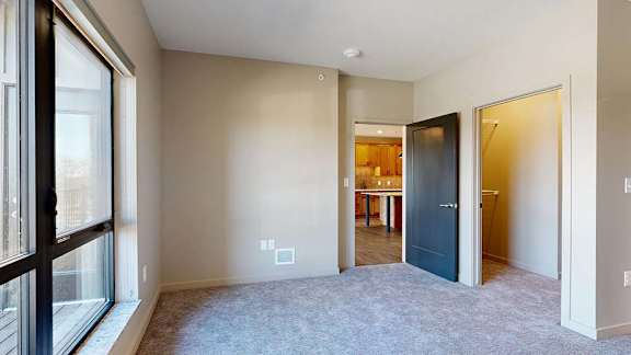 Bright apartment bedroom with neutral carpeting, a large window for natural light, and an open doorway leading to the adjacent living area and kitchen.