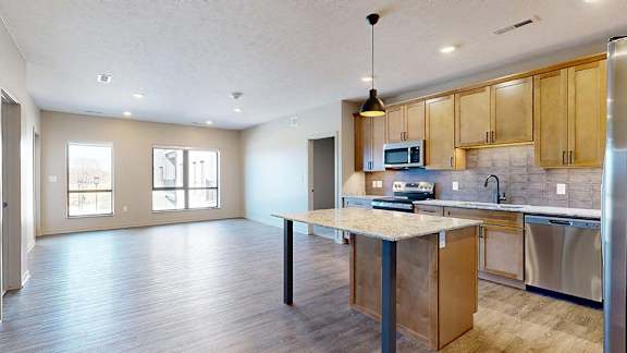 Bright open-concept apartment kitchen and living area with a center island, wood cabinetry, stainless steel appliances, pendant lighting, and large windows.